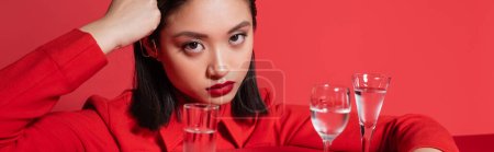 brunette asian woman in jacket looking at camera near glasses with clear water isolated on red, banner