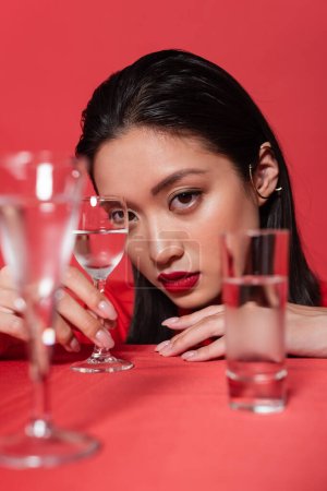 Porträt einer asiatischen Frau mit Make-up und Ohrenmanschette in der Nähe von Gläsern mit klarem Wasser auf verschwommenem Vordergrund isoliert auf Rot
