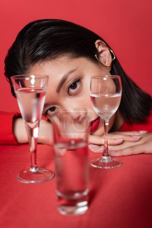 portrait of brunette asian woman with ear cuff and makeup looking at camera near blurred glasses with water isolated on red