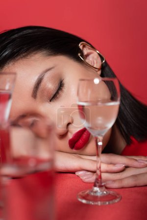 portrait of asian woman with makeup and closed eyes near glasses with pure water on blurred foreground isolated on red