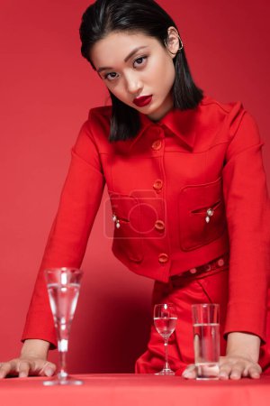 brunette asian woman in elegant jacket standing near glasses with clean water on red background