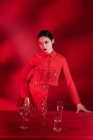 mujer asiática de moda con maquillaje artístico mirando a la cámara cerca de gafas con agua clara sobre fondo rojo con sombra