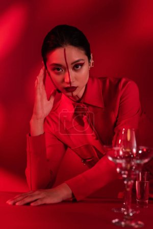 Téléchargez les photos : Femme asiatique avec visage créatif posant dans une veste à la mode près de verres flous avec de l'eau sur fond rouge ombragé - en image libre de droit