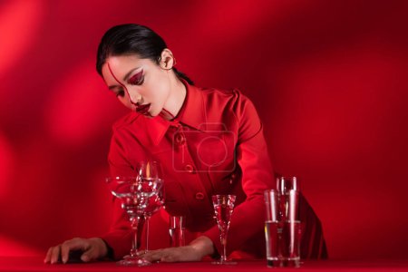Téléchargez les photos : Femme asiatique avec maquillage artistique debout dans une veste à la mode près de différents verres avec de l'eau sur fond rouge - en image libre de droit