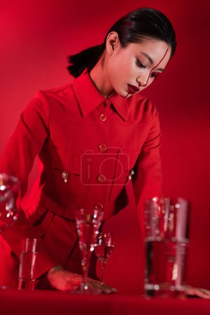 morena mujer asiática en chaqueta de moda y maquillaje creativo de pie cerca de gafas borrosas sobre fondo rojo