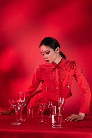 brünette asiatische Frau in trendigem Outfit und kreativem Gesicht neben verschiedenen Gläsern mit Wasser auf rotem abstrakten Hintergrund