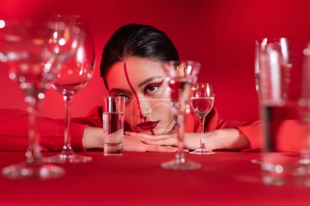 junge asiatische Frau mit kreativem Make-up im Gesicht geteilt mit Linie in der Nähe verschwommene Gläser Wasser auf rotem Hintergrund