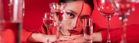 Téléchargez les photos : Jeune femme avec maquillage artistique regardant la caméra près de verres d'eau flous isolés sur rouge, bannière - en image libre de droit