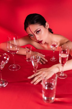 naked asian woman with creative makeup posing near different glasses with water on red background