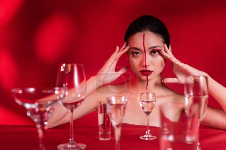 asian woman with bare shoulders and artistic makeup looking at camera near blurred glasses with water on red background