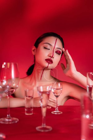 Téléchargez les photos : Brunette asiatique femme avec les yeux fermés et le visage artistique près de verres flous sur fond rouge - en image libre de droit