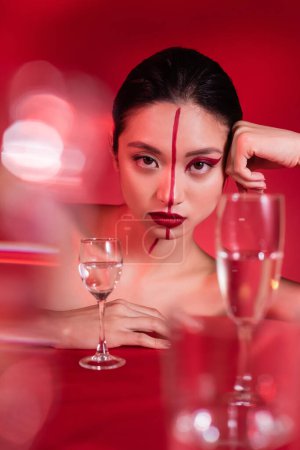 Téléchargez les photos : Portrait de femme asiatique avec maquillage sur le visage divisé avec ligne regardant la caméra près de lunettes floues sur fond rouge - en image libre de droit