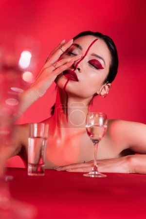 asian woman with closed eyes touching face with creative makeup near blurred glasses on red background