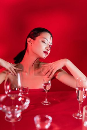 Téléchargez les photos : Sensuelle asiatique femme avec les yeux fermés et créatif visage touchant épaules nues près de verres flous sur fond rouge - en image libre de droit