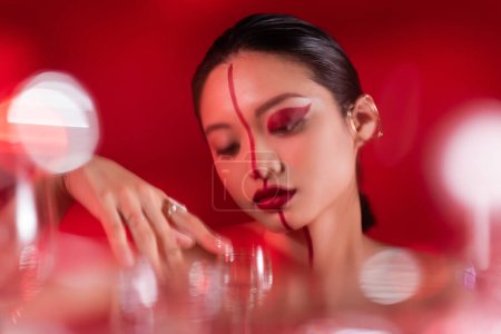 brünette asiatische Frau mit künstlerischem Gesicht in der Nähe einer verschwommenen Brille auf rotem Hintergrund
