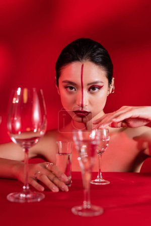 nude asian woman with creative makeup and line on face touching glass with pure water on red background