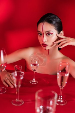 asian woman with bright creative visage and hand near face looking at camera near various glasses on red background