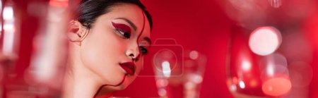 Porträt einer asiatischen Frau mit kreativem Make-up im Gesicht geteilt durch eine Linie in der Nähe einer verschwommenen Brille auf rotem Hintergrund, Banner
