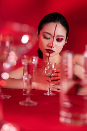 young asian woman with red artistic visage looking at glass with pure water on blurred foreground 
