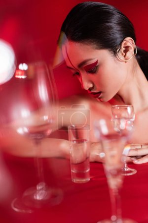 Foto de Desnudo asiático mujer con oreja manguito y creativo visage cerca borrosa vasos de agua en rojo fondo - Imagen libre de derechos
