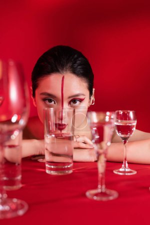 sinnliche asiatische Frau mit artistischem Make-up blickt in die Kamera nahe Gläser mit reinem Wasser auf rotem Hintergrund