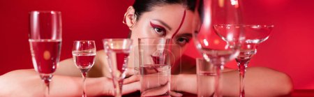 Téléchargez les photos : Asiatique femme avec artistique visage et oreille poignets regarder caméra près de lunettes avec pure eau isolé sur rouge, bannière - en image libre de droit