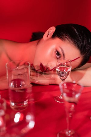 brünette asiatische Frau mit artistischem Make-up liegt und schaut in die Kamera in der Nähe einer verschwommenen Brille auf rotem Hintergrund