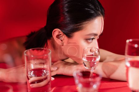 brünette asiatische Frau mit Make-up sieht weg in der Nähe verschwommene Gläser mit reinem Wasser auf rotem Hintergrund