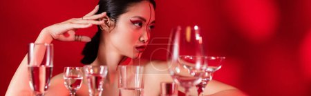 Téléchargez les photos : Brunette asiatique femme avec des épaules nues et visage créatif regardant loin près de différents verres sur fond rouge, bannière - en image libre de droit