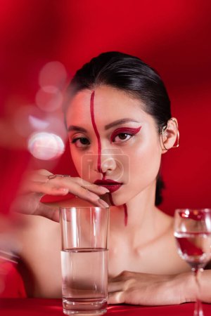 Porträt einer asiatischen Frau mit artistischem Make-up im Gesicht geteilt durch eine Linie, die Glas mit reinem Wasser auf rotem Hintergrund berührt