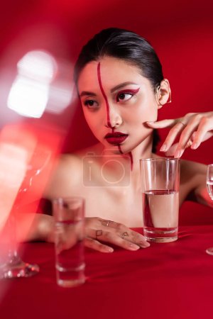 nude asian woman with artistic makeup and ear cuff looking away near blurred glasses on red background