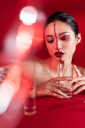 nude asian woman with red artistic makeup holding glass of clear water on blurred foreground