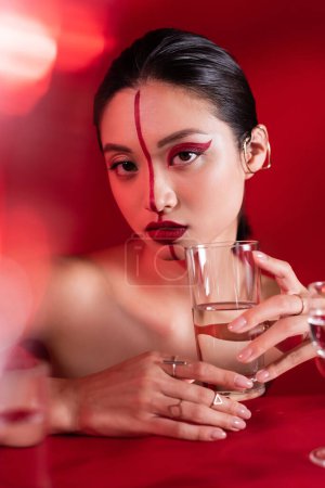 nude asian woman with red creative makeup holding glass of water and looking at camera on blurred foreground