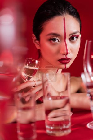 Porträt einer charmanten asiatischen Frau mit kreativem Gesicht, die in die Kamera schaut, nahe einer verschwommenen Brille auf rotem Hintergrund
