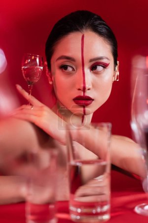 Porträt einer jungen Asiatin mit rotem künstlerischem Make-up, die Glas mit klarem Wasser auf verschwommenem Vordergrund hält
