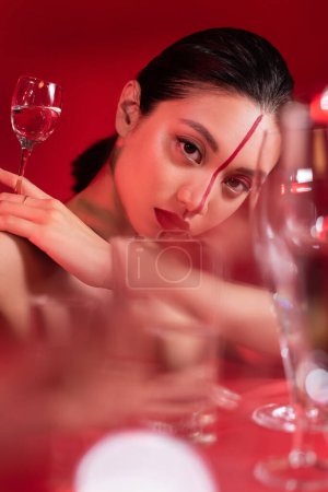 Téléchargez les photos : Portrait de femme asiatique sensuelle avec visage artistique regardant la caméra près de lunettes floues sur fond rouge - en image libre de droit