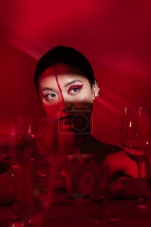 Téléchargez les photos : Jeune femme asiatique avec maquillage artistique regardant loin dans la lumière près de lunettes floues sur fond rouge foncé - en image libre de droit