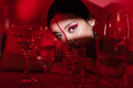 asian woman with artistic makeup and ear cuff looking at camera in light near blurred glasses on dark red background