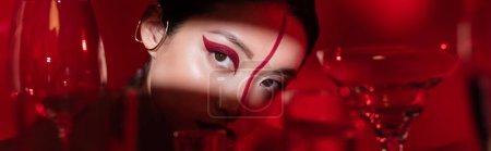 charming asian woman with bright artistic makeup looking away in light near blurred glasses on red background, banner