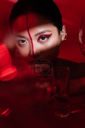 Foto de Mujer asiática con rostro creativo y manguito de oído mirando a la cámara en la luz cerca de gafas borrosas sobre fondo rojo oscuro - Imagen libre de derechos