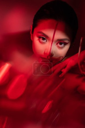 Porträt einer asiatischen Frau mit artistischem Make-up, die auf rotem, verschwommenem Vordergrund in die Kamera blickt