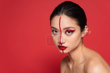 sexy asian woman with bare shoulders posing in artistic makeup and stylish ear cuff isolated on red