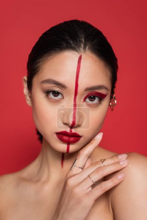 portrait of brunette asian woman with hand near face and creative visage looking at camera isolated on red
