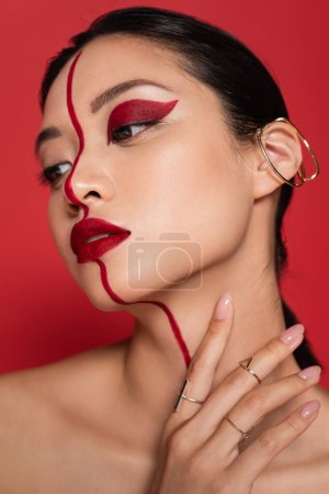 retrato de morena mujer asiática con maquillaje creativo y arete de puño sosteniendo la mano cerca del cuello aislado en rojo