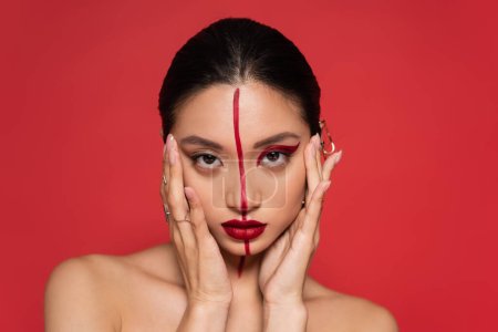 brunette asian woman with bare shoulders and artistic makeup holding hands near face isolated on red