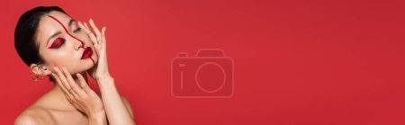Photo for Sensual asian woman with bare shoulders and artistic visage touching perfect face with closed eyes isolated on red, banner - Royalty Free Image