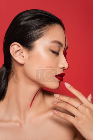 Profil einer asiatischen Frau mit geschlossenen Augen und kreativer Visage, die die Hand in Gesichtsnähe hält, isoliert auf Rot