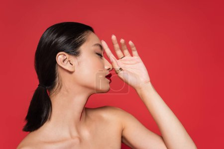 perfil de morena mujer asiática con hombros desnudos y maquillaje posando con la mano cerca de la cara aislada en rojo