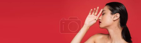 Photo for Profile of sexy asian woman with bare shoulder and bright visage holding hand near face isolated on red, banner - Royalty Free Image