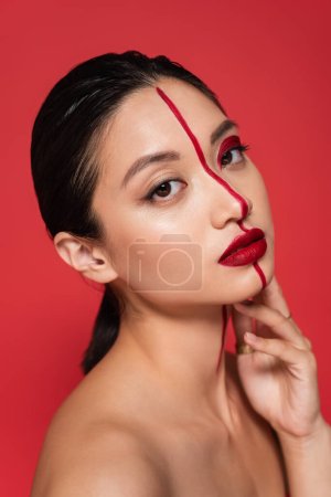 retrato de modelo asiático con rostro creativo y piel perfecta cogida de la mano cerca de la cara y mirando a la cámara aislada en rojo